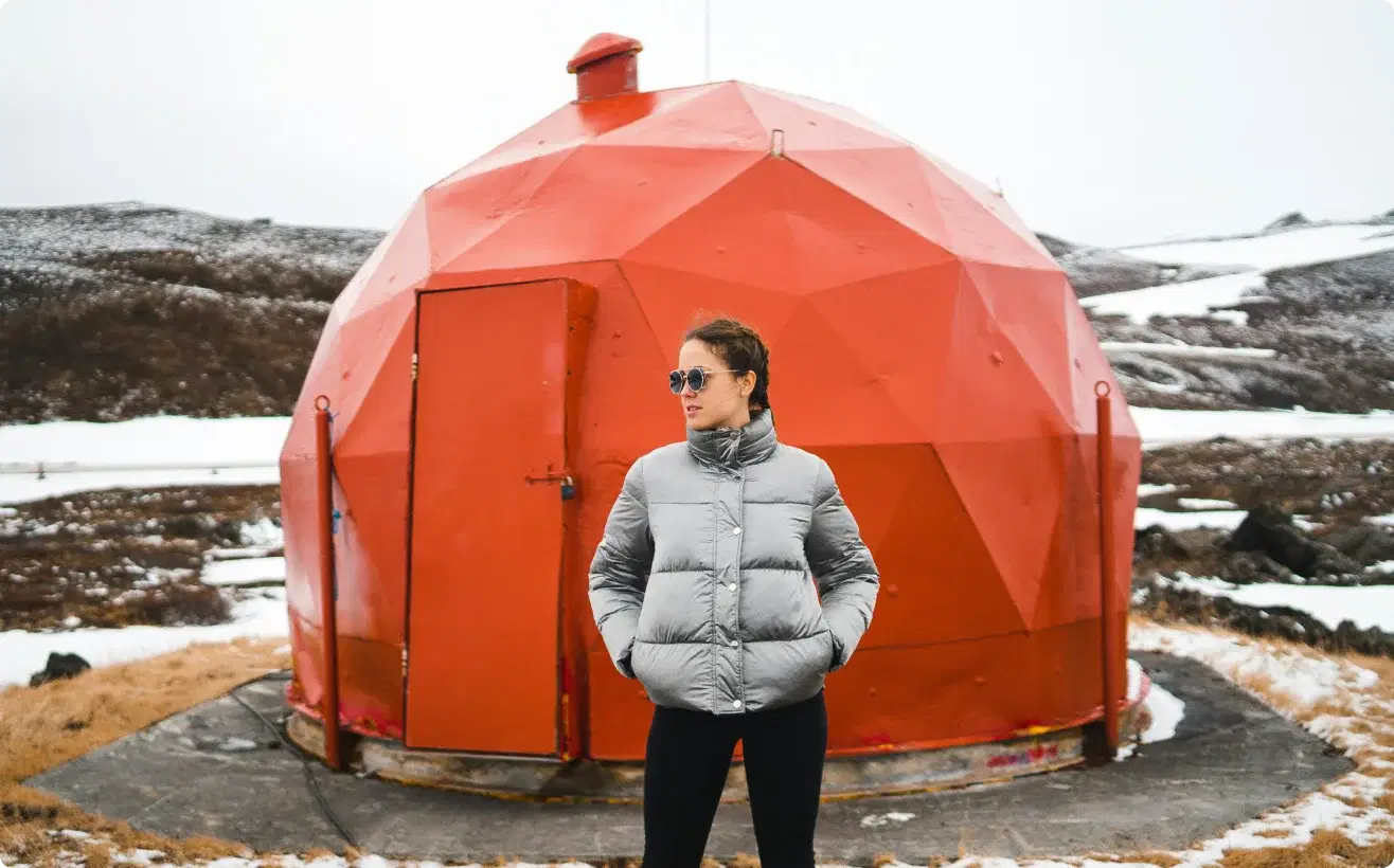 Person in a winter jacket standing in front of a unique red geodesic structure in Iceland, symbolizing adventure and the opportunity to join the Cars Iceland Affiliate Program.