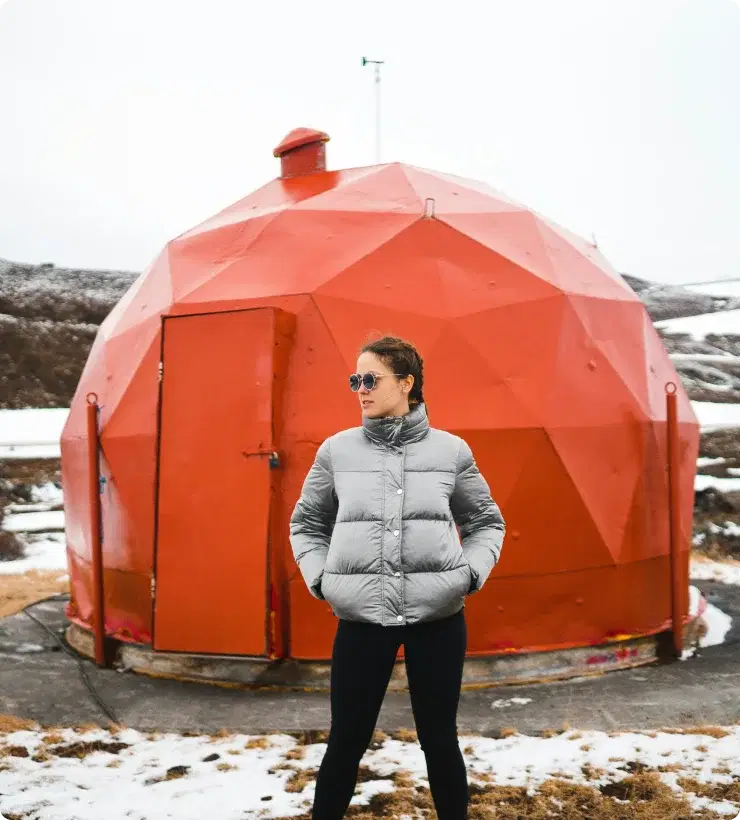 Person in a winter jacket standing in front of a unique red geodesic structure in Iceland, symbolizing adventure and the opportunity to join the Cars Iceland Affiliate Program.