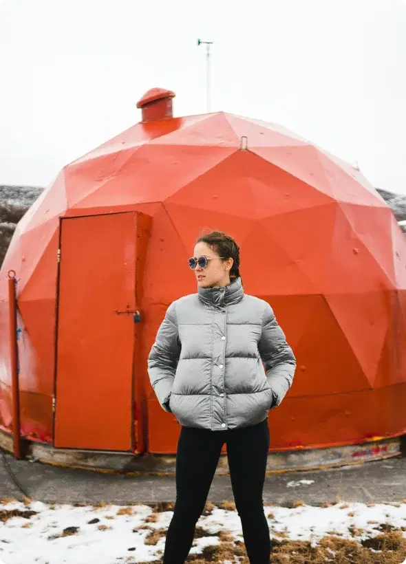 Person in a winter jacket standing in front of a unique red geodesic structure in Iceland, symbolizing adventure and the opportunity to join the Cars Iceland Affiliate Program.