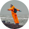 Man in bright orange winter gear standing on rocky terrain with arms outstretched.