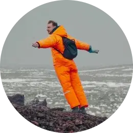 Man in bright orange winter gear standing on rocky terrain with arms outstretched.