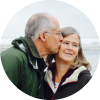 Elderly man kissing a woman on the cheek with an outdoor background.