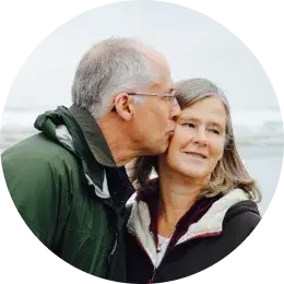 Elderly man kissing a woman on the cheek with an outdoor background.