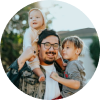 Smiling man carrying two young children on his shoulders outdoors.