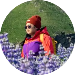 Traveler in a red beanie and colorful jacket walking through a field of purple lupines in Iceland.