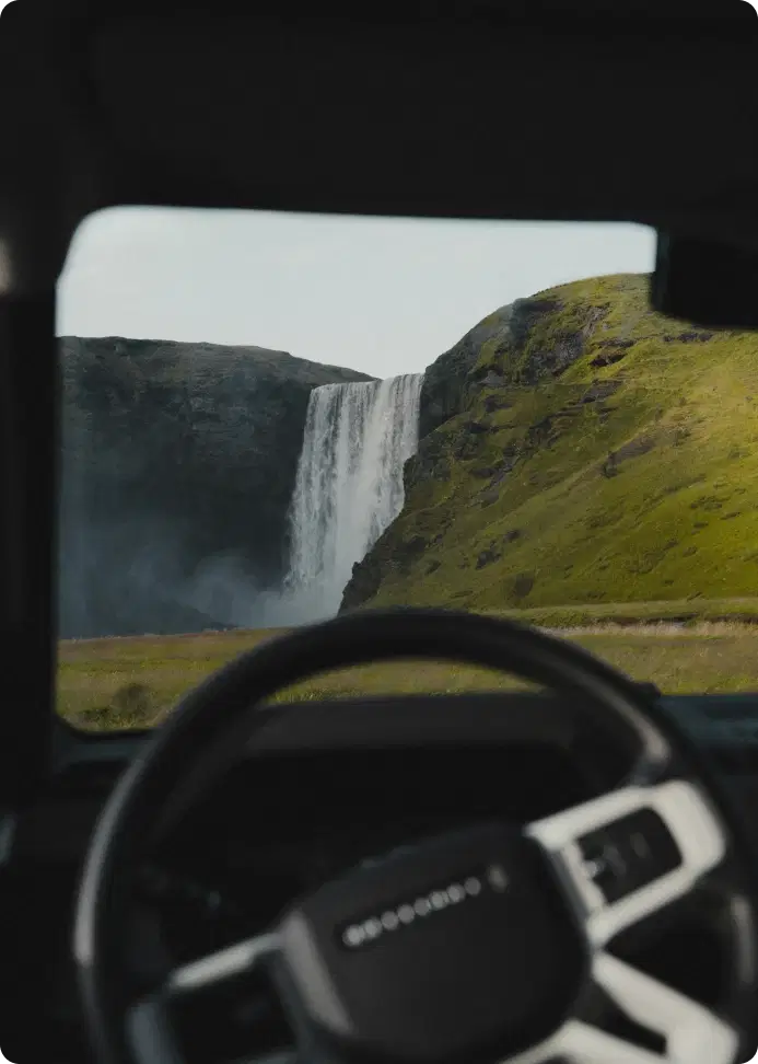 View of Skógafoss waterfall through a car windshield in Iceland, capturing the iconic waterfall framed by the vehicle’s steering wheel.