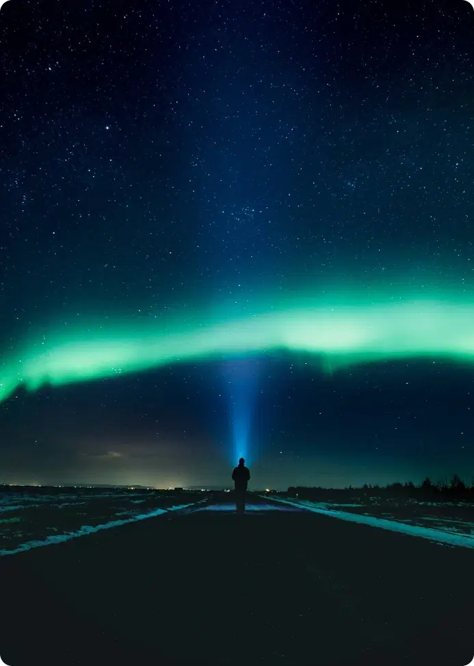 Silhouette of a person with a flashlight under the Northern Lights in Iceland, illuminating the night sky filled with stars.