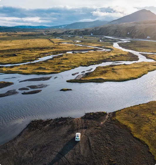 A lone 4x4 SUV parked near a river in Iceland's vast, open wilderness, surrounded by winding waterways, grassy plains, and distant mountains under a cloudy sky.