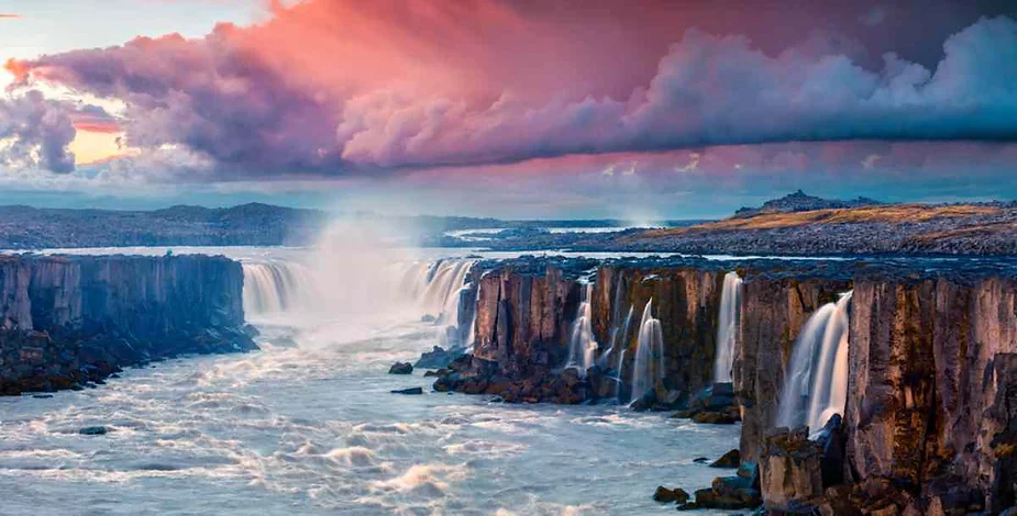 Breathtaking view of a powerful Icelandic waterfall flowing through a dramatic canyon with pink and purple clouds during sunset, showcasing nature's beauty