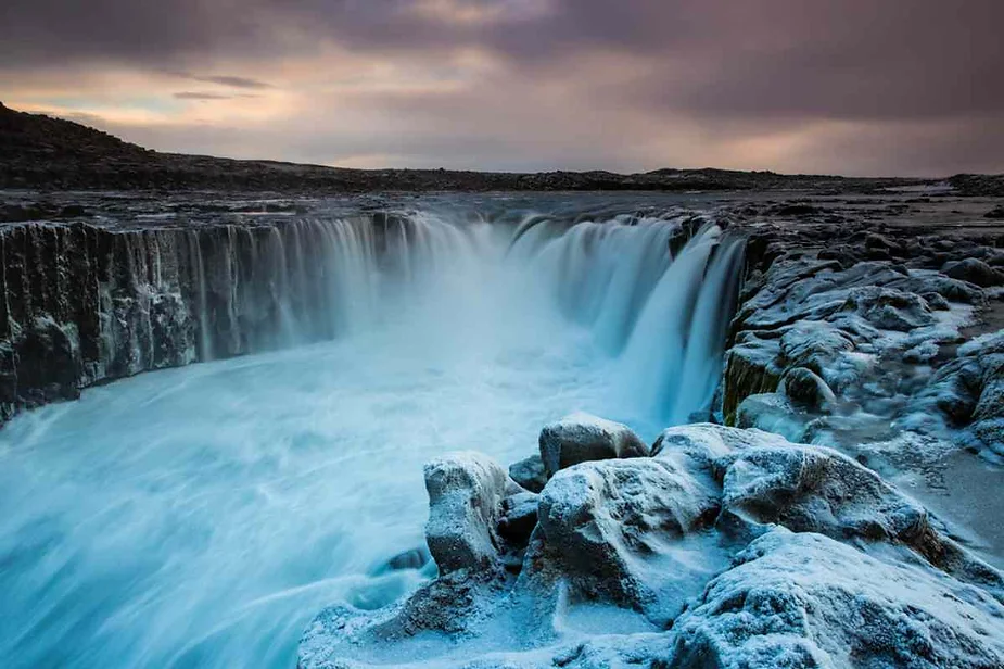 Majestic waterfall in Iceland cascading over basalt columns into a frothy blue pool, surrounded by rugged, icy terrain under a dramatic sky