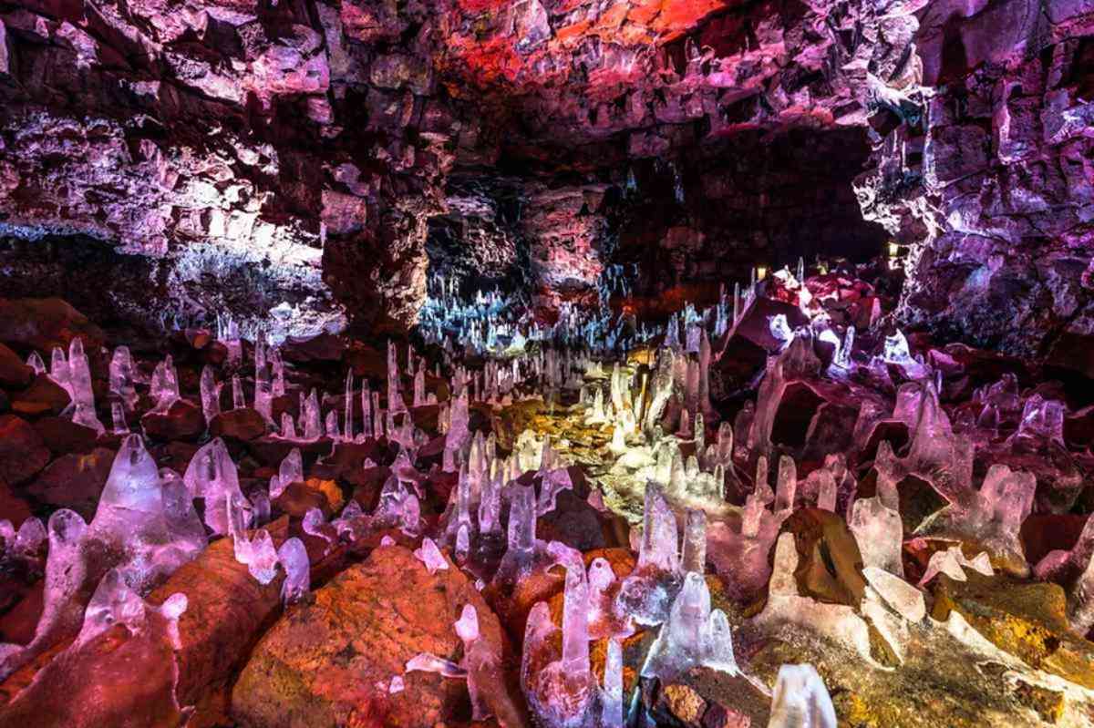 Interior of an Icelandic lava cave with numerous ice stalagmites illuminated by colorful lights, showcasing a stunning contrast between ice and rock formations