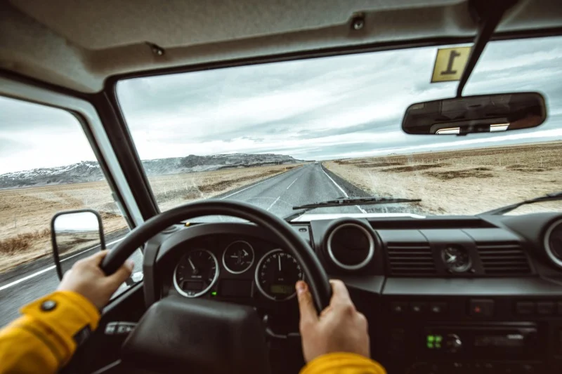 View from inside a car, with the driver holding the steering wheel, driving on a long, empty road through a barren landscape.