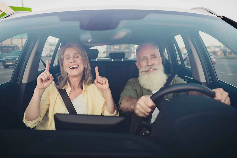 A cheerful older couple in a car, with the woman happily raising her hands and the man smiling while driving.