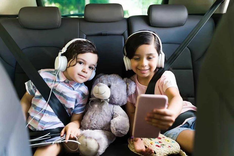 Two children sitting in the back seat of a car, wearing headphones, watching something on a smartphone, with a stuffed toy bear between them.