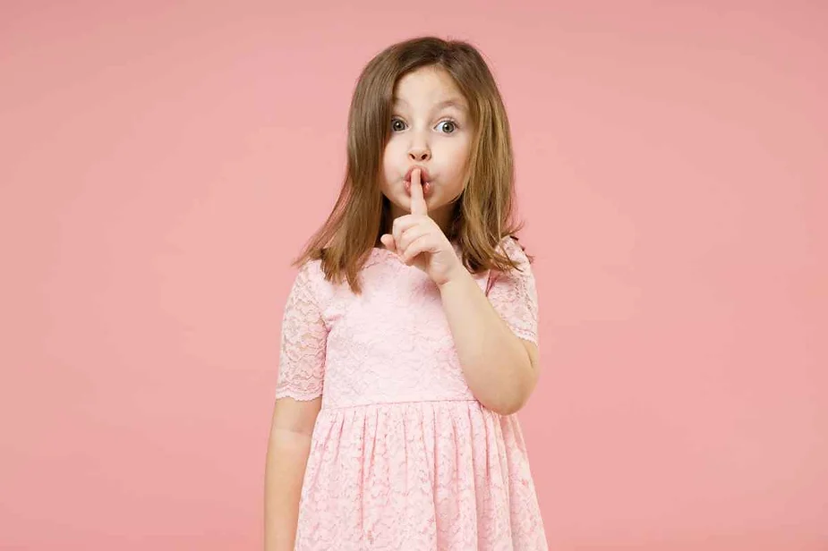 A young girl in a pink dress holding her finger to her lips, making a 