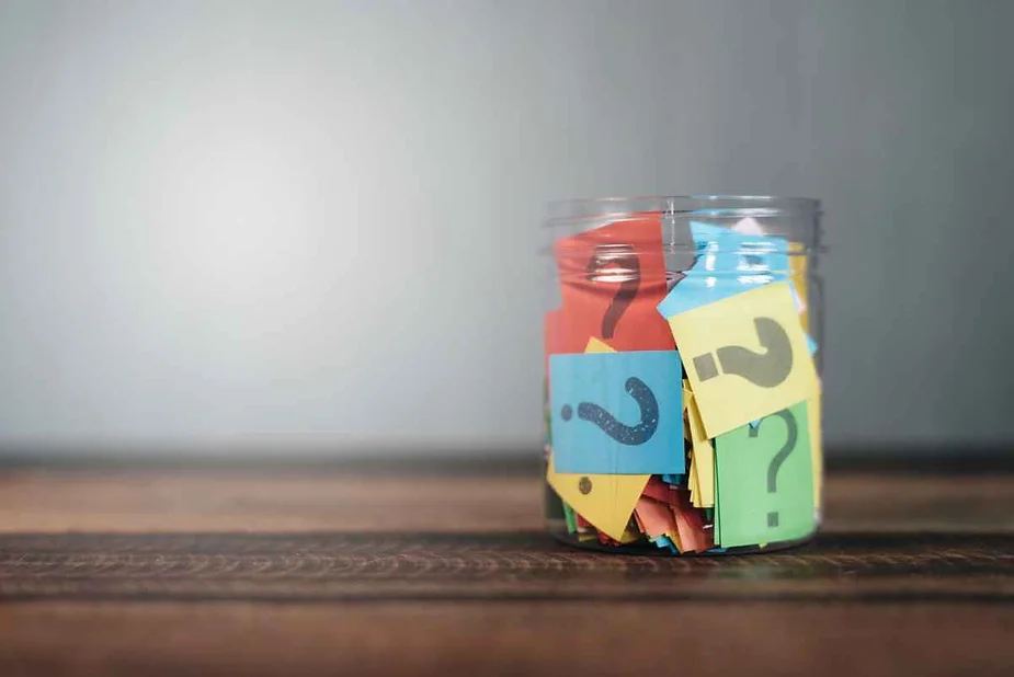 A clear jar filled with colorful paper slips, each marked with a question mark, sitting on a wooden surface.