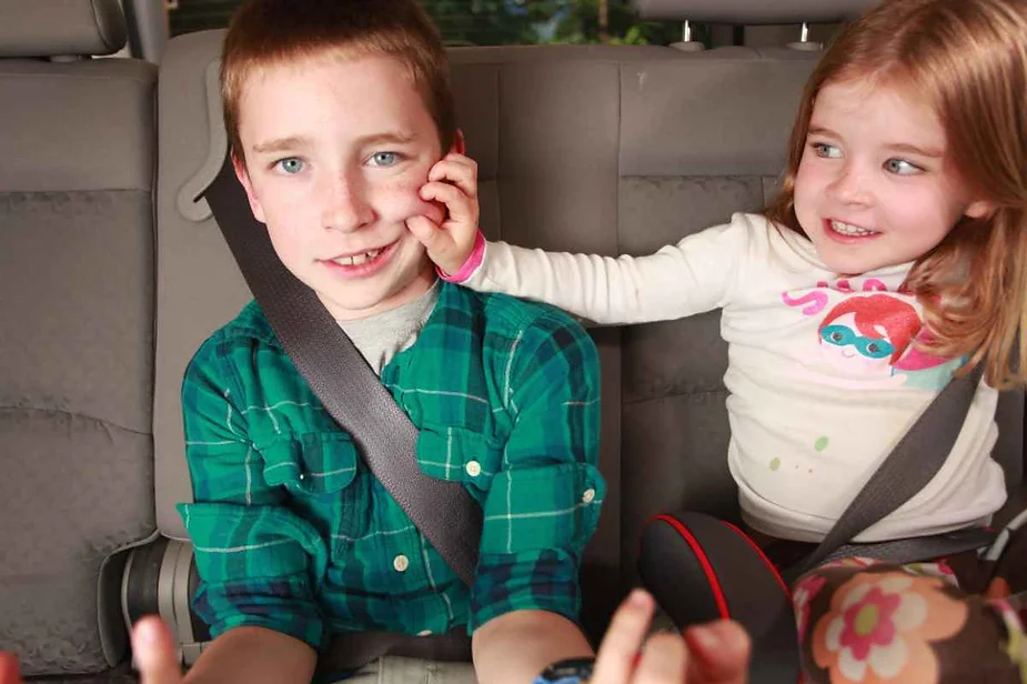 Two young children sitting in the back seat of a car, with the girl playfully pinching the boy's cheek, both wearing seatbelts.