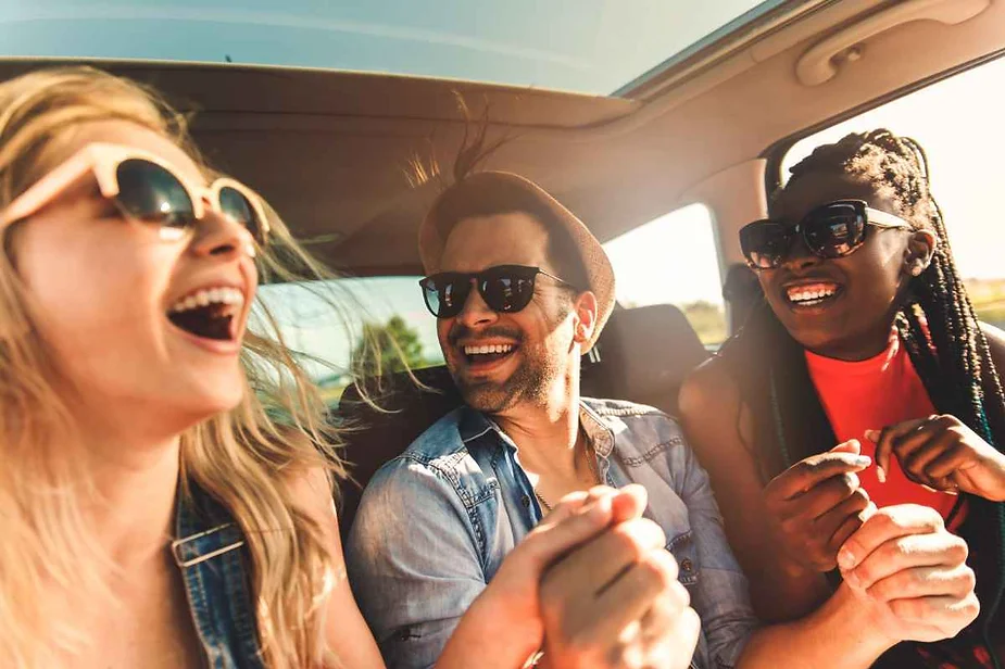 Three friends wearing sunglasses, smiling and laughing together in a car during a road trip, enjoying a sunny day.