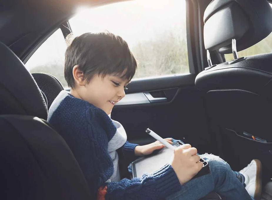 A young boy sitting in the backseat of a car, smiling while drawing on a tablet, enjoying his time during a journey.