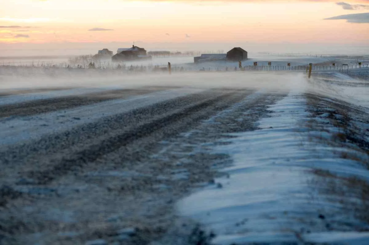 A winter scene in Iceland with a snow-covered road and houses in the distance, under a colorful sunset sky.