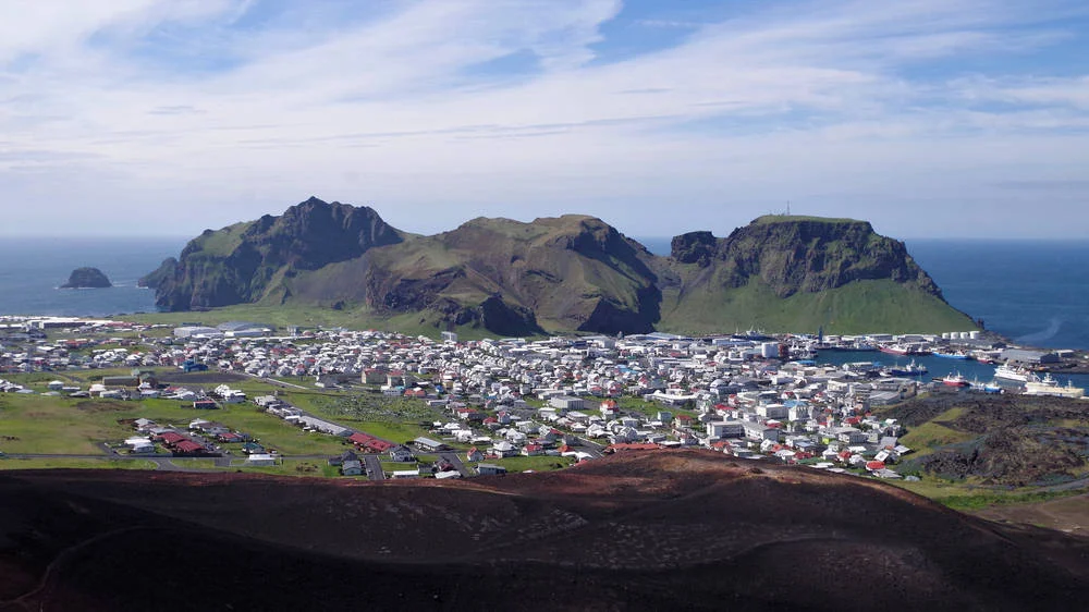 An aerial view of the town of Heimaey on Heimaey Island in Iceland, with the Eldfell Volcano and its surrounding green mountains in the background.