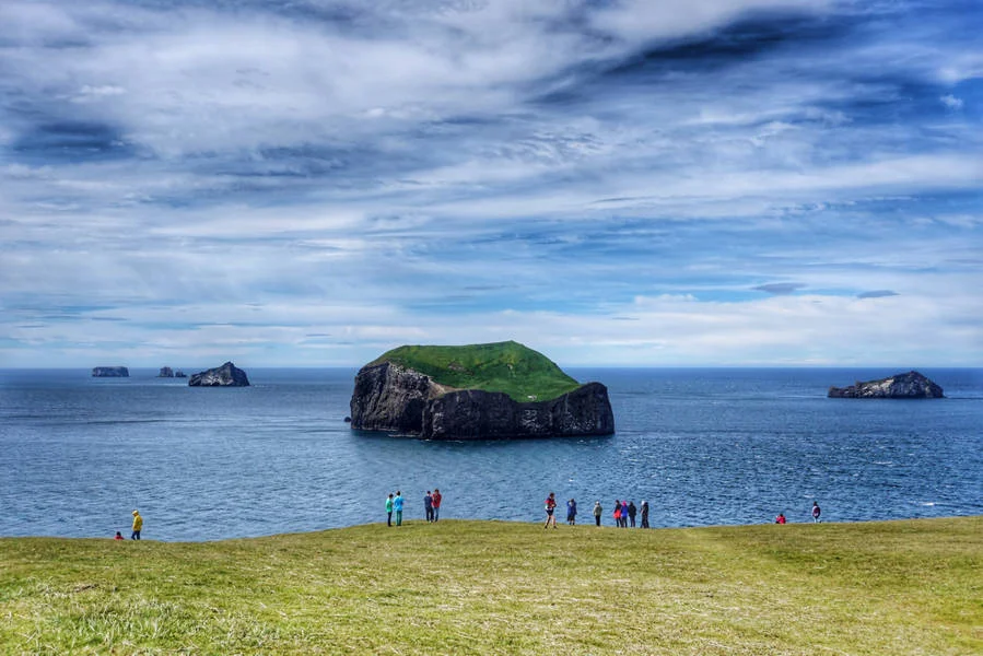 A group of people stands on a grassy hill overlooking the ocean with the Westman Islands, characterized by their rocky formations and green tops, visible in the distance under a partly cloudy sky.