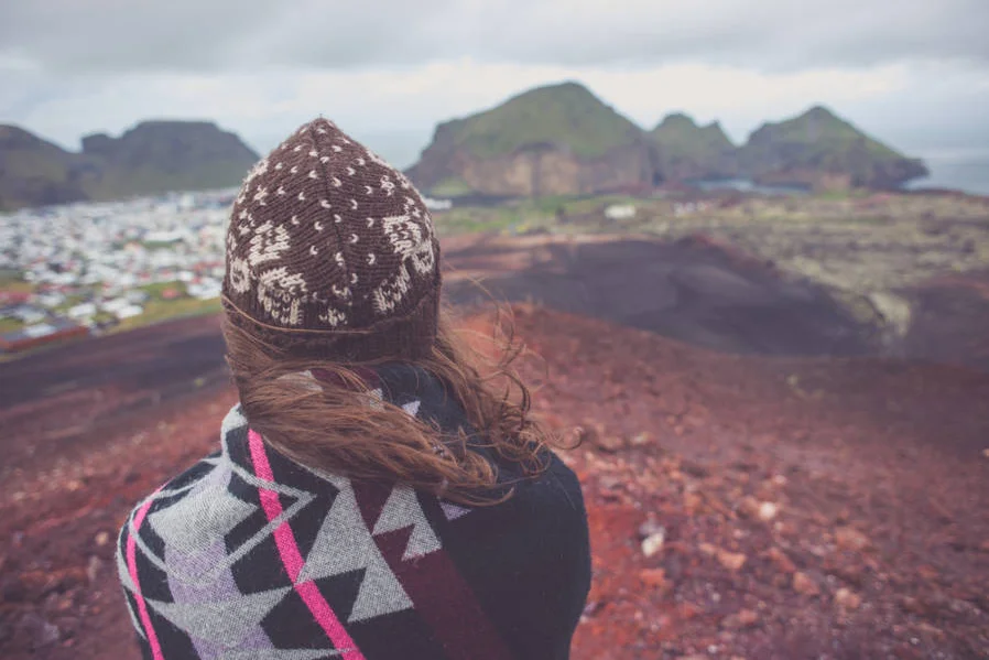 A woman wearing a knitted hat and a patterned blanket stands on the red slopes of Eldfell Volcano, overlooking the town of Heimaey and the surrounding green mountains on Heimaey Island, Iceland.