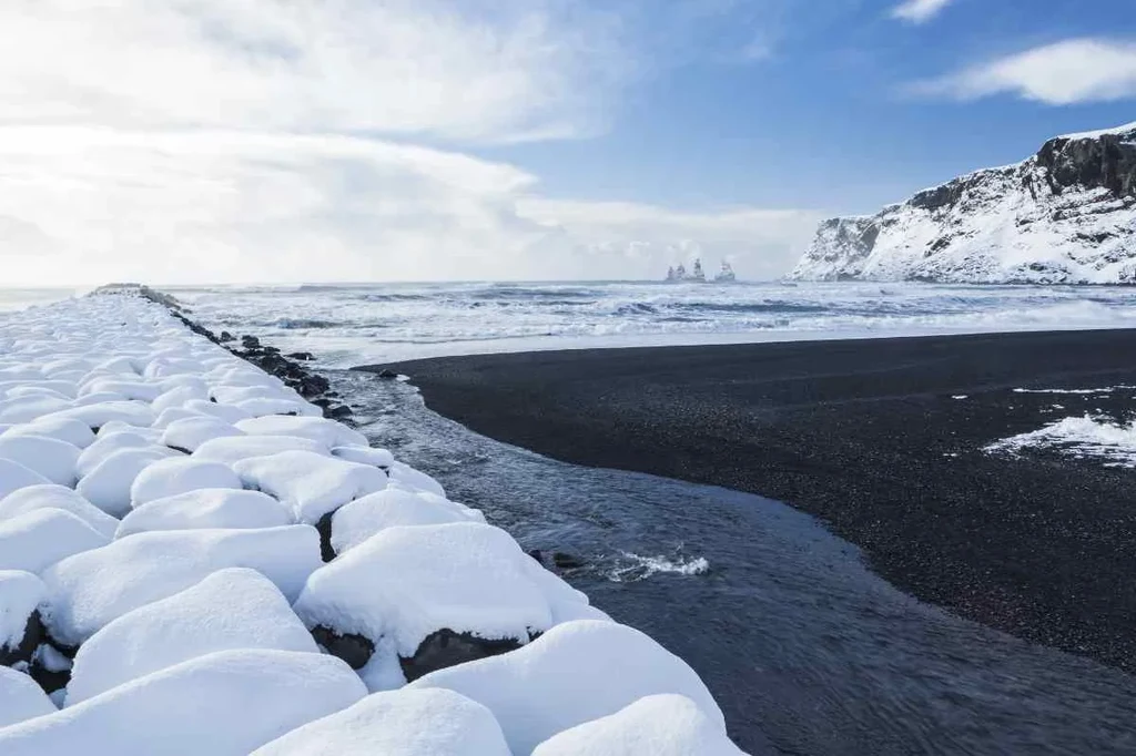 Snow-covered rocks on the shore of a black sand beach in Iceland, with waves crashing and distant sea stacks under a bright blue sky, highlighting the contrast between snow and volcanic sand.