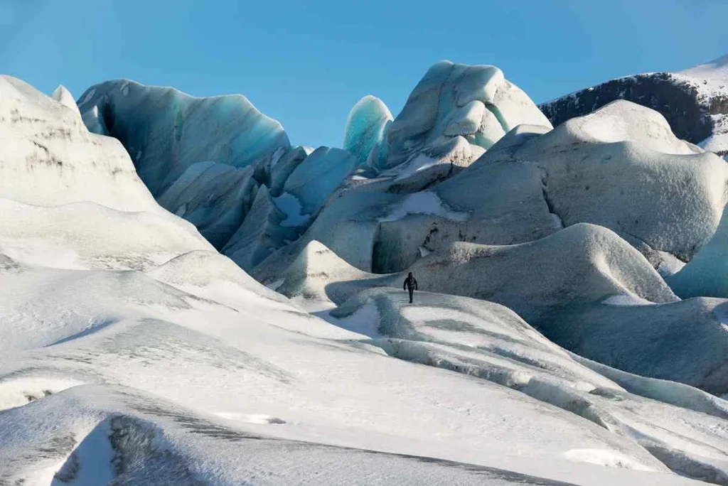 A person walking across a vast, icy landscape with towering glaciers in the background under a clear blue sky in Iceland, highlighting the awe-inspiring beauty and scale of the glacial formations.