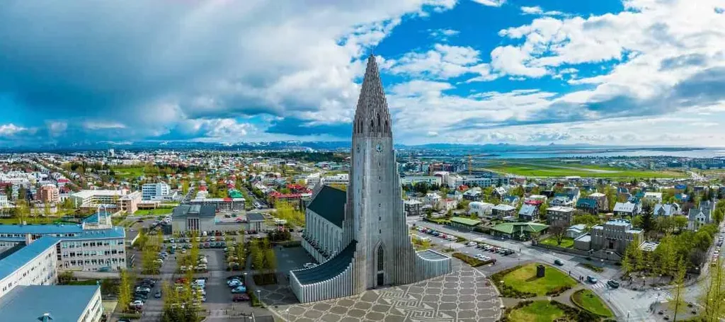 Aerial view of the Hallgrimskirkja church in Reykjavik, Iceland, with its distinctive tall spire and modern design, surrounded by the colorful cityscape under a bright blue sky.