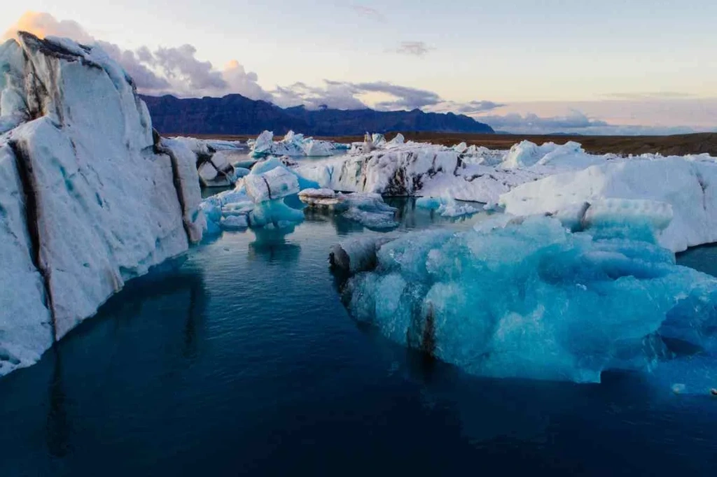 Large icebergs floating in the serene waters of Jökulsárlón Glacier Lagoon in Iceland, with a backdrop of mountains and a partly cloudy sky, highlighting the stunning natural beauty and icy landscape.