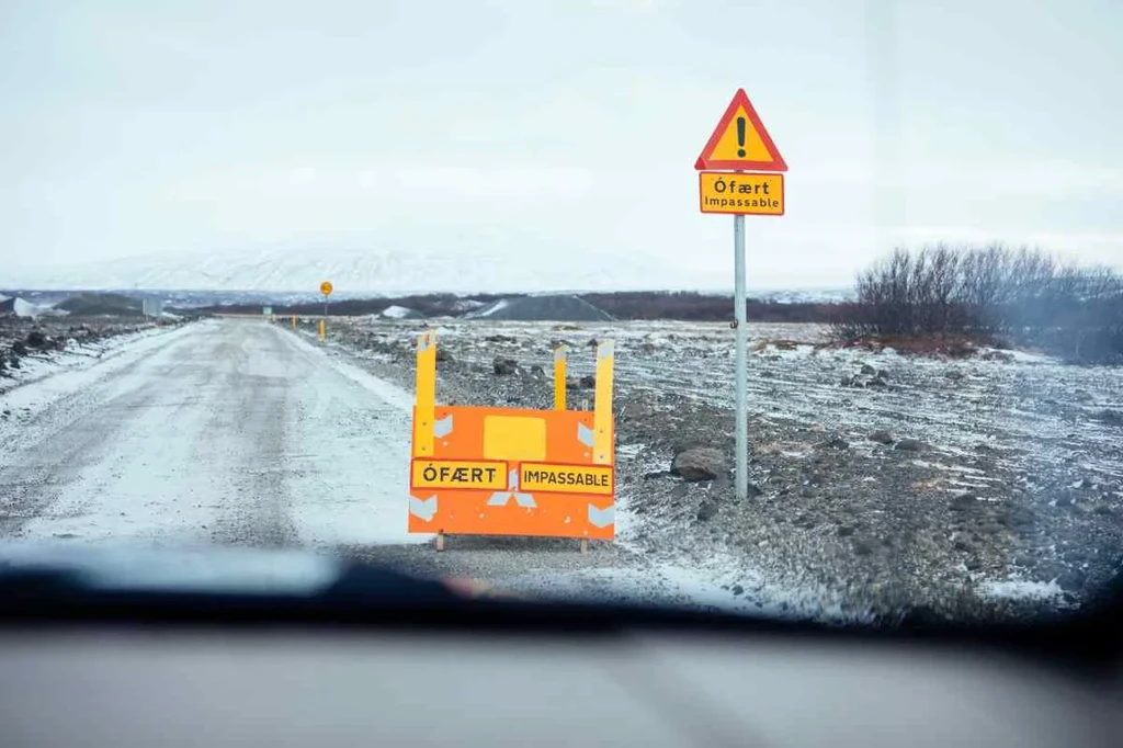 A snow-covered road in Iceland with an orange and yellow road sign indicating 'impassable due to severe winter conditions' viewed from inside a vehicle