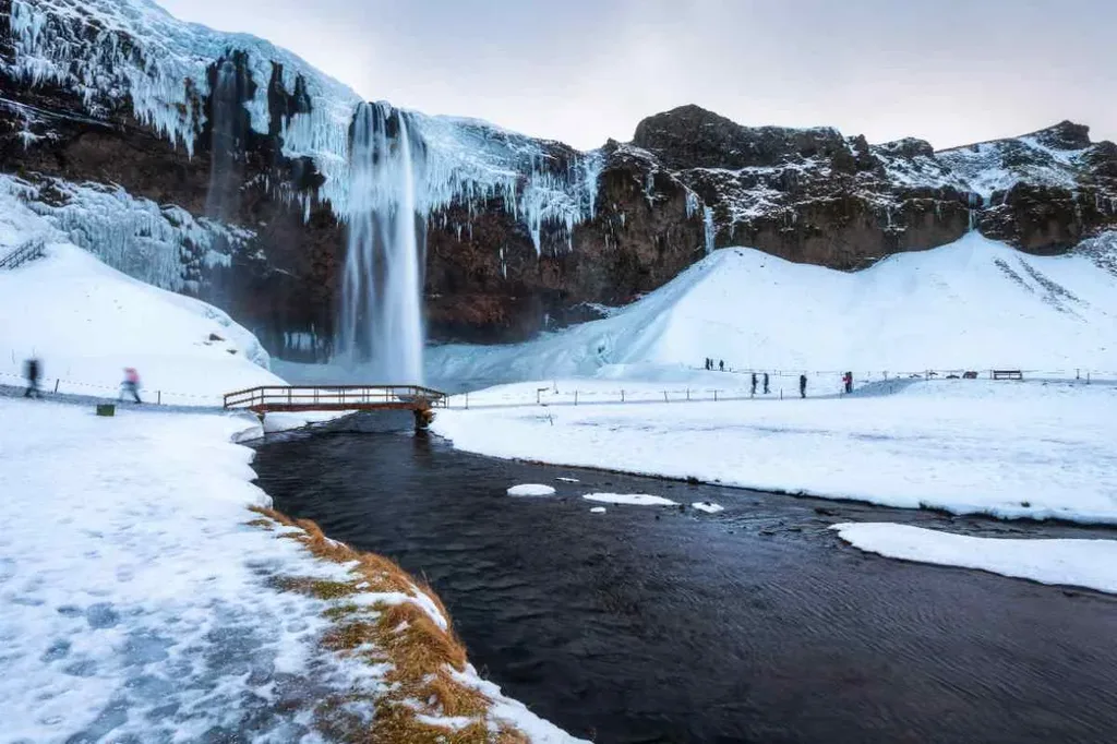 Seljalandsfoss waterfall in Iceland during winter, with icicles hanging from the cliffs, a snow-covered landscape, and a small bridge over a stream, showcasing the enchanting winter scenery.