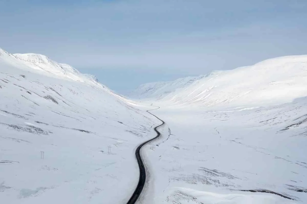 A winding road cuts through a vast, snow-covered valley flanked by snowy mountains under a clear sky in Iceland during winter, showcasing the serene and remote landscape.