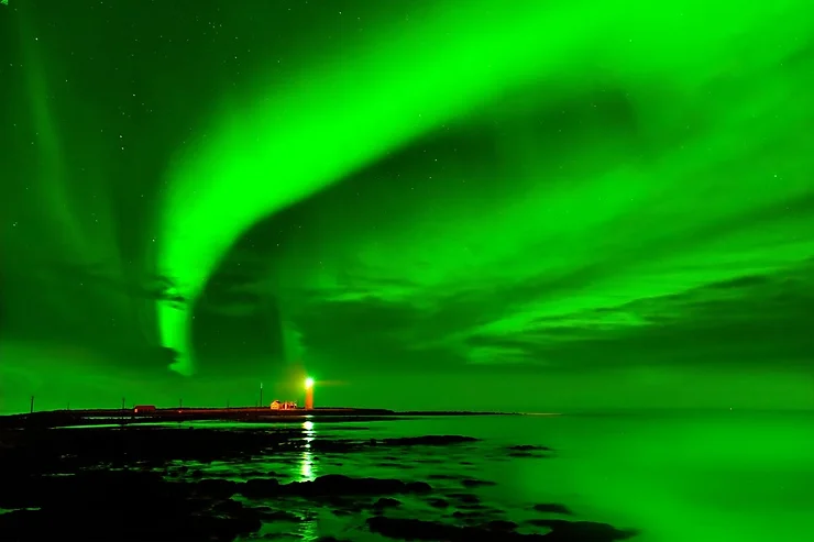 Vivid green Northern Lights dance in the night sky above Grotta Lighthouse, reflecting off the calm waters below, creating a breathtaking and serene spectacle.