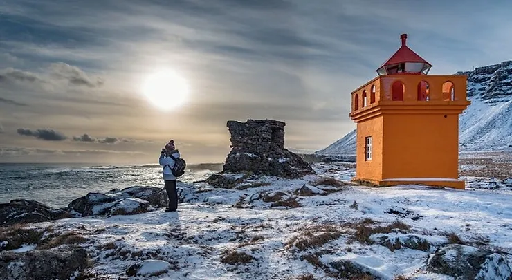 A person stands near the bright orange Hafnarnesviti Lighthouse on a snowy, rocky landscape, with the sun casting a soft glow over the scene.