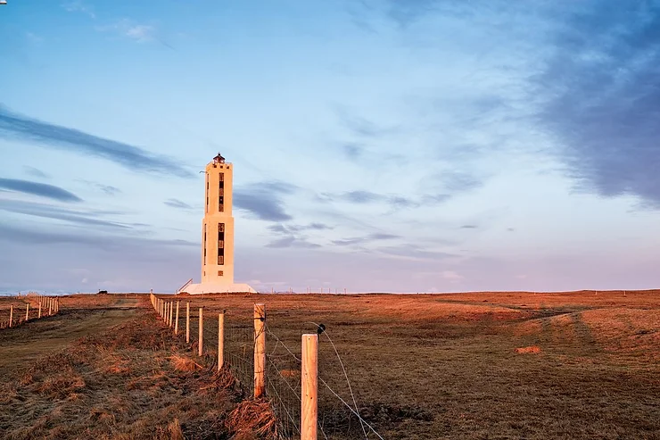 The Knarrarósviti Lighthouse stands tall on an open field, bathed in the warm hues of a setting sun, with a clear sky overhead.