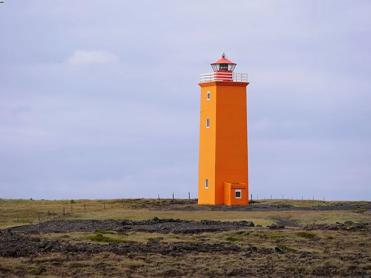 The vibrant orange Selvogsviti Lighthouse stands tall on a flat, grassy plain in Iceland, its bright color contrasting with the overcast sky.