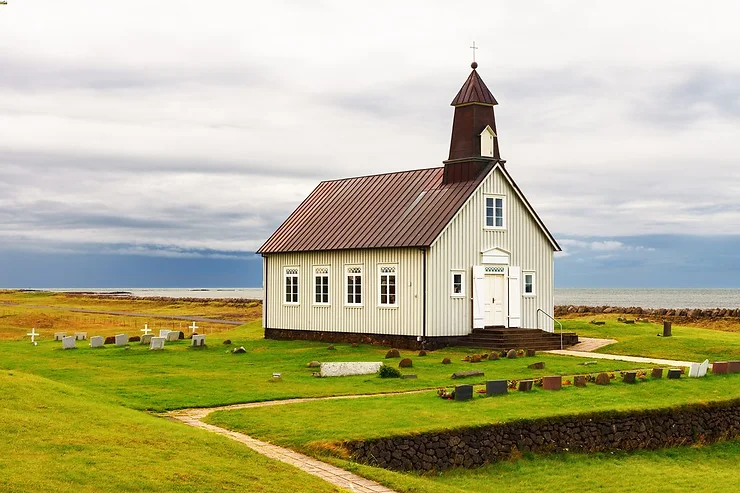 Strandar Church, a charming wooden church with a brown roof, stands surrounded by lush green grass and a serene graveyard, under a cloudy sky with the ocean in the background.