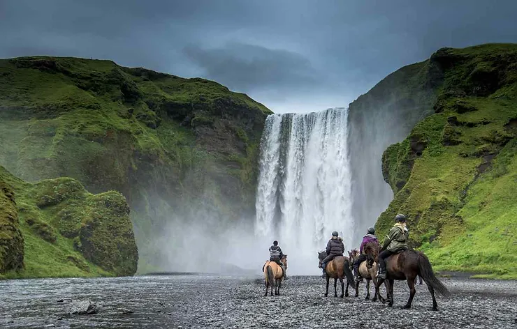 A group of riders on Icelandic horses approaches a majestic waterfall, surrounded by lush green cliffs. The mist from the waterfall adds a mystical touch to the scene, epitomizing Iceland's rugged and beautiful landscape.