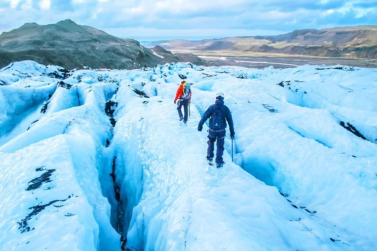 Two people equipped with winter hiking gear traverse a vast glacier with deep crevasses. The icy blue surface contrasts with the distant rugged, brown mountains under a clear sky, showcasing Iceland's stunning winter landscape.