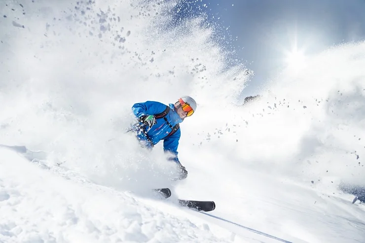 A skier in a bright blue jacket and helmet speeds down a snowy slope, creating a spray of powdery snow. The sun shines brightly in the background, illuminating the exhilarating scene of a winter skiing vacation.