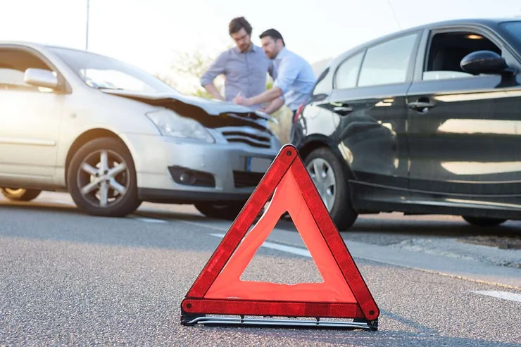 A red warning triangle in the foreground with two men examining the damage to their cars after a collision in the background. One car is silver, and the other is black, illustrating the aftermath of a car accident.