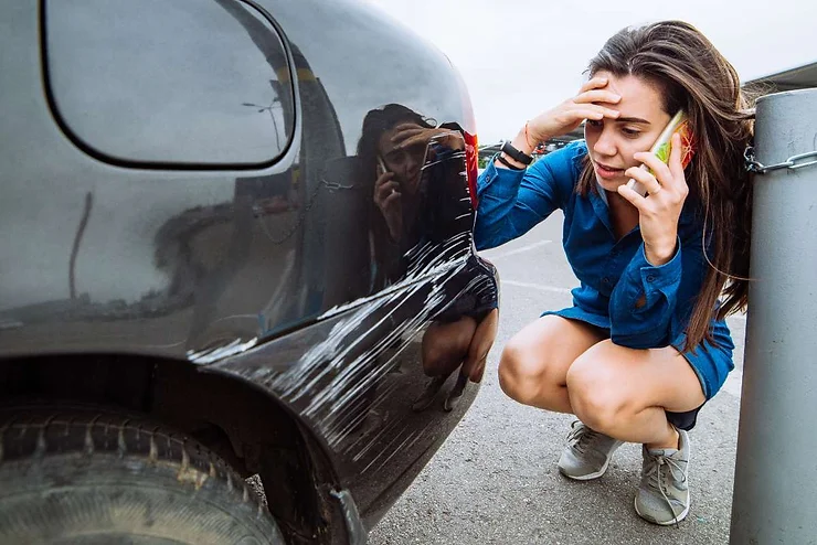 A distressed woman kneeling beside a scratched black rental car, holding a phone to her ear while examining the damage.