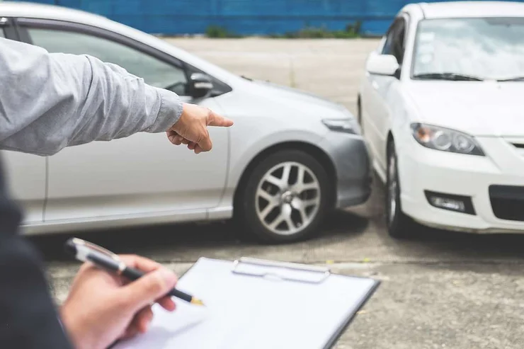 A person holding a clipboard in the foreground while another individual points at the damage between a silver and a white car after a minor collision.