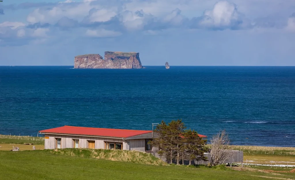 Scenic view of Drangey Island off the coast of Iceland, with a red-roofed building and lush green fields in the foreground, showcasing the serene and picturesque landscape.