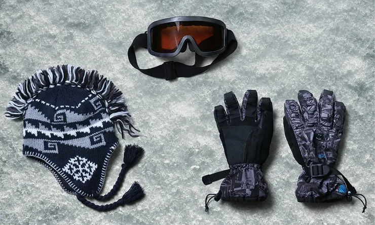 A set of essential gear for glacier hiking tours, including a warm patterned hat with tassels, protective goggles, and a pair of insulated gloves, all laid out on a snowy surface.