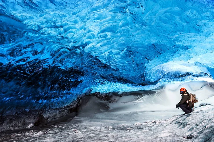 A person wearing a helmet and backpack crouches inside a stunning blue ice cave, surrounded by glistening icy walls and a snowy floor.