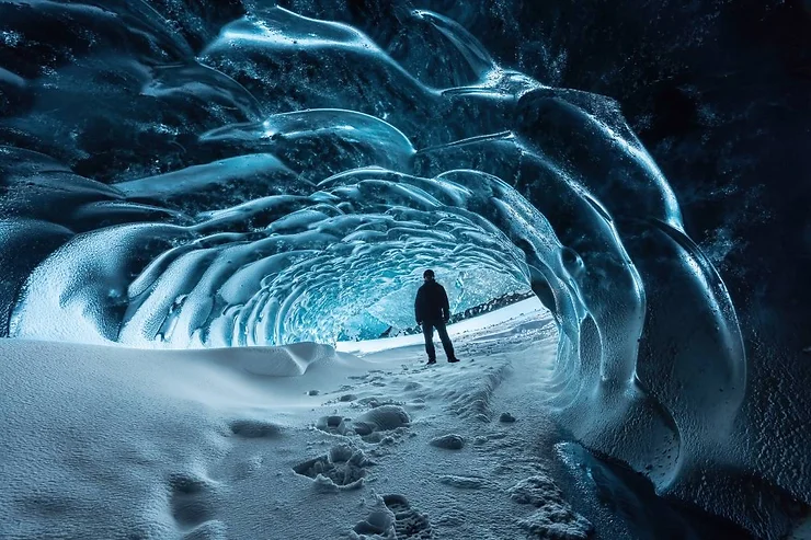 A person stands inside a mesmerizing ice tunnel in Iceland, surrounded by icy blue walls and a snowy floor, creating a breathtaking and surreal landscape.