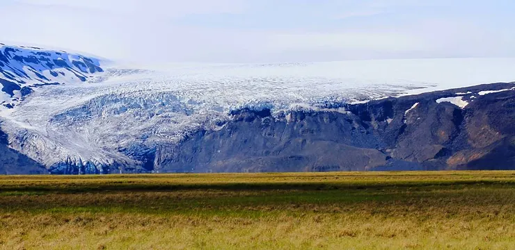 A sweeping view of Katla Glacier in Iceland, showcasing its expansive ice formations cascading down from the mountain, surrounded by a vast, grassy plain under a clear sky.